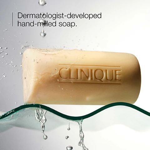 Image 1 - A bar of hand-milled soap with the word CLINIQUE embossed on it is shown surrounded by droplets of water. Dermatologist-developed hand-milled soap.
Image 2 - Two bars of Clinique facial soap are displayed: one in a light yellow color labeled Clinique Facial Soap - Dry, and the other in a light beige color labeled Clinique Facial Soap - Oily, with both having descriptions for their skin type.