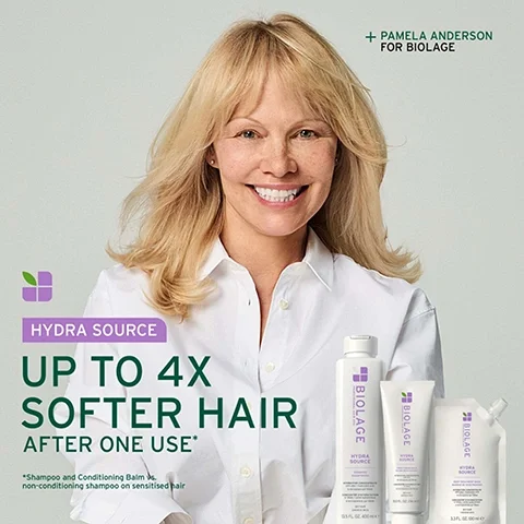 Image 2 - A smiling blonde woman, Pamela Anderson, wearing a white shirt, is shown with three Biolage Hydra Source hair products including a shampoo, a conditioning balm, and a deep treatment mask, with visible text that reads + PAMELA ANDERSON FOR BIOLAGE, HYDRA SOURCE, UP TO 4X SOFTER HAIR AFTER ONE USE*, and *Shampoo and Conditioning Balm vs. non-conditioning shampoo on sensitised hair.